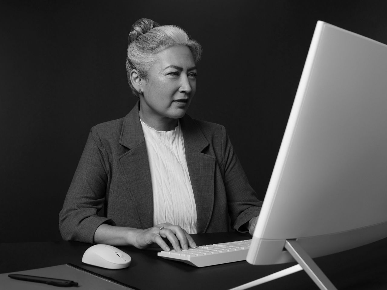 It is an image of a female office administrator seated at her desk while viewing a monitor in an office.