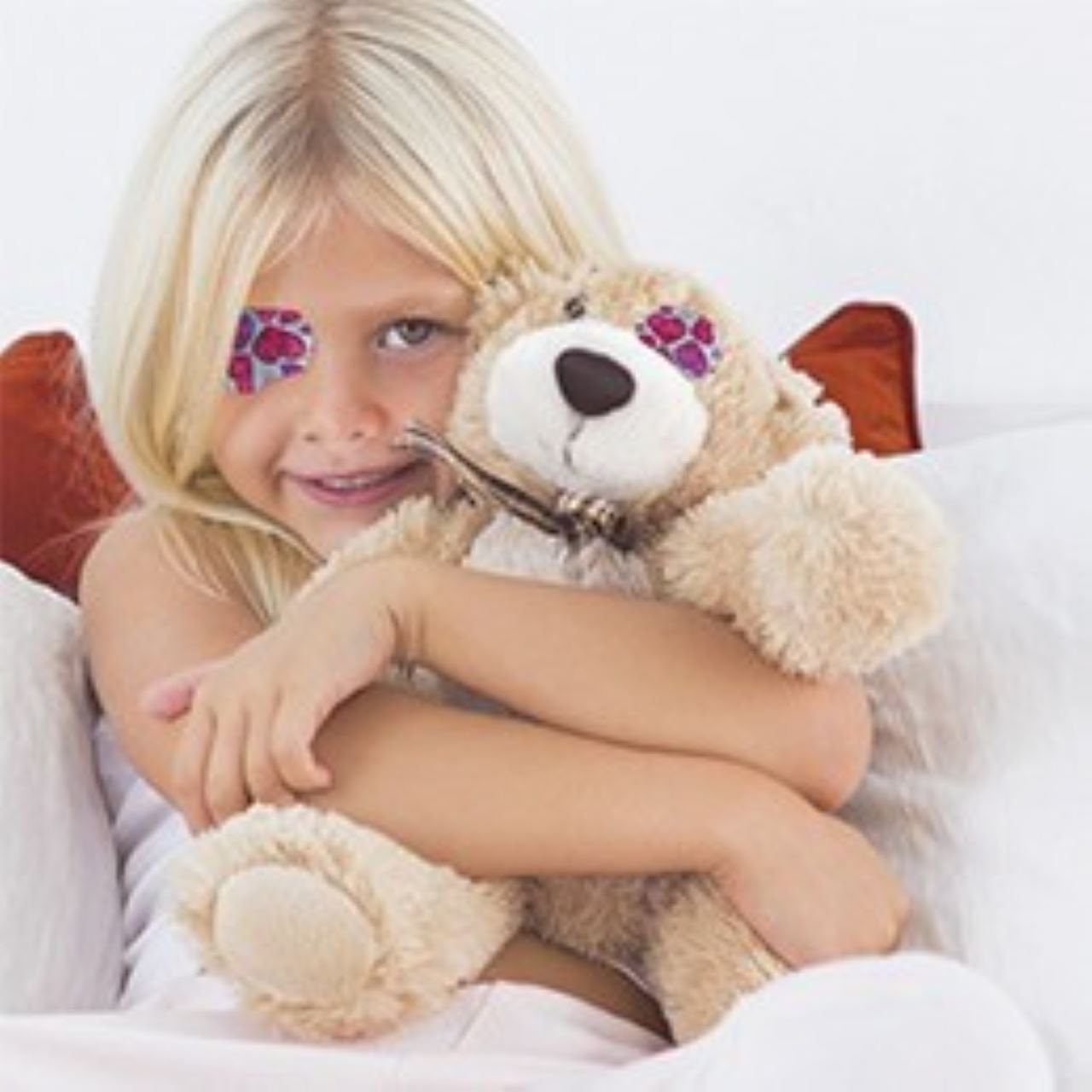 Blond pige med Opticlude silikone øjenplaster med hjerte motiv, smilende og krammende en beige og hvid bamse