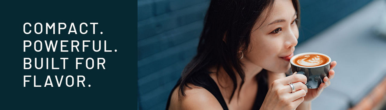 Woman drinking a cup of cappachino in a coffee shop with text that reads Compact. Powerful. Built for Flavor, which are the benefits of the HRFO system.