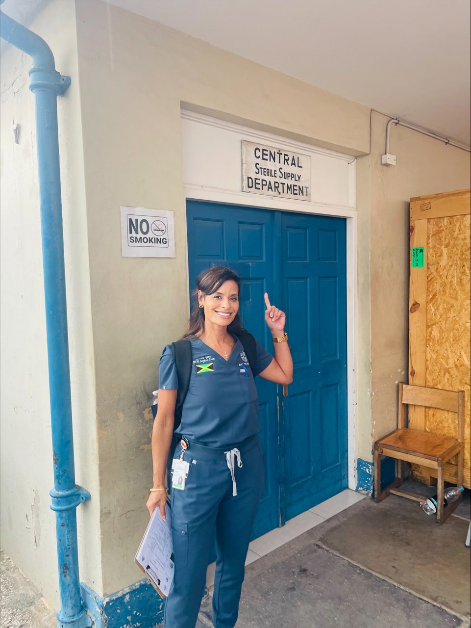 Senovia Gunn, dressed in scrubs and smiling, stands outside the Central Sterile Supply Department in Jamaica, holding a clipboard.