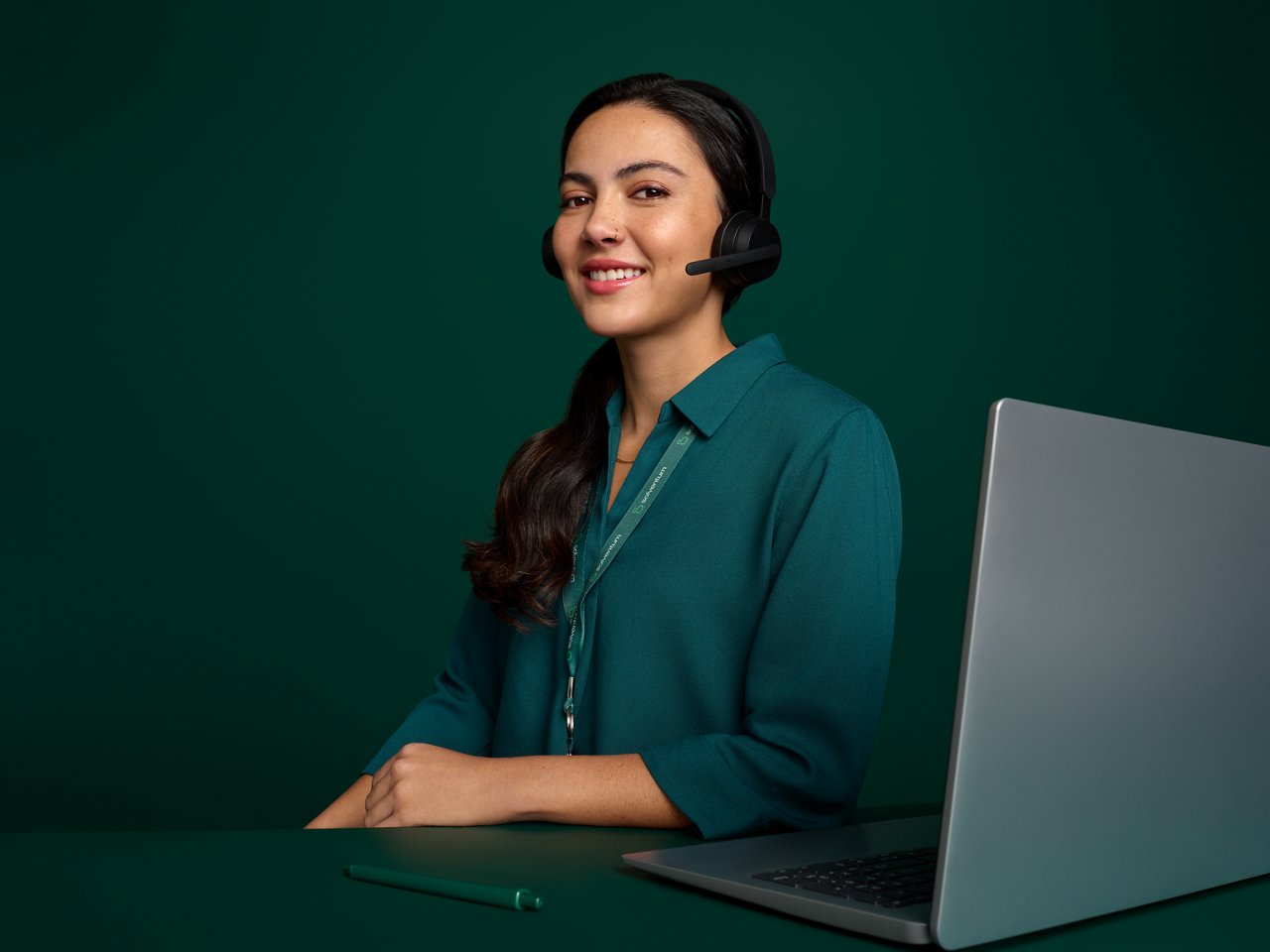 Portrait of a female call center employee smiling, seated at her desk from the Costa Rica office on a dark green background.