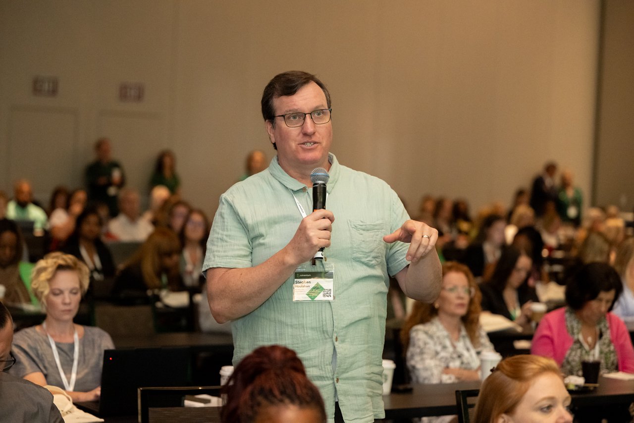 Man with short brown hair, wearing a green shirt and lanyard, standing in audience, holding and speaking into a microphone.