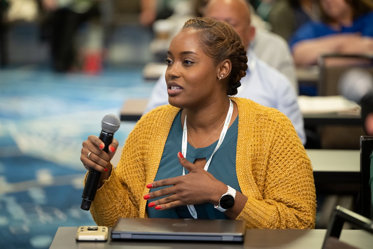 Woman seated at the 2025 Solventum CES, wearing a teal top and yellow sweater, holding and sepaking into a microphone.