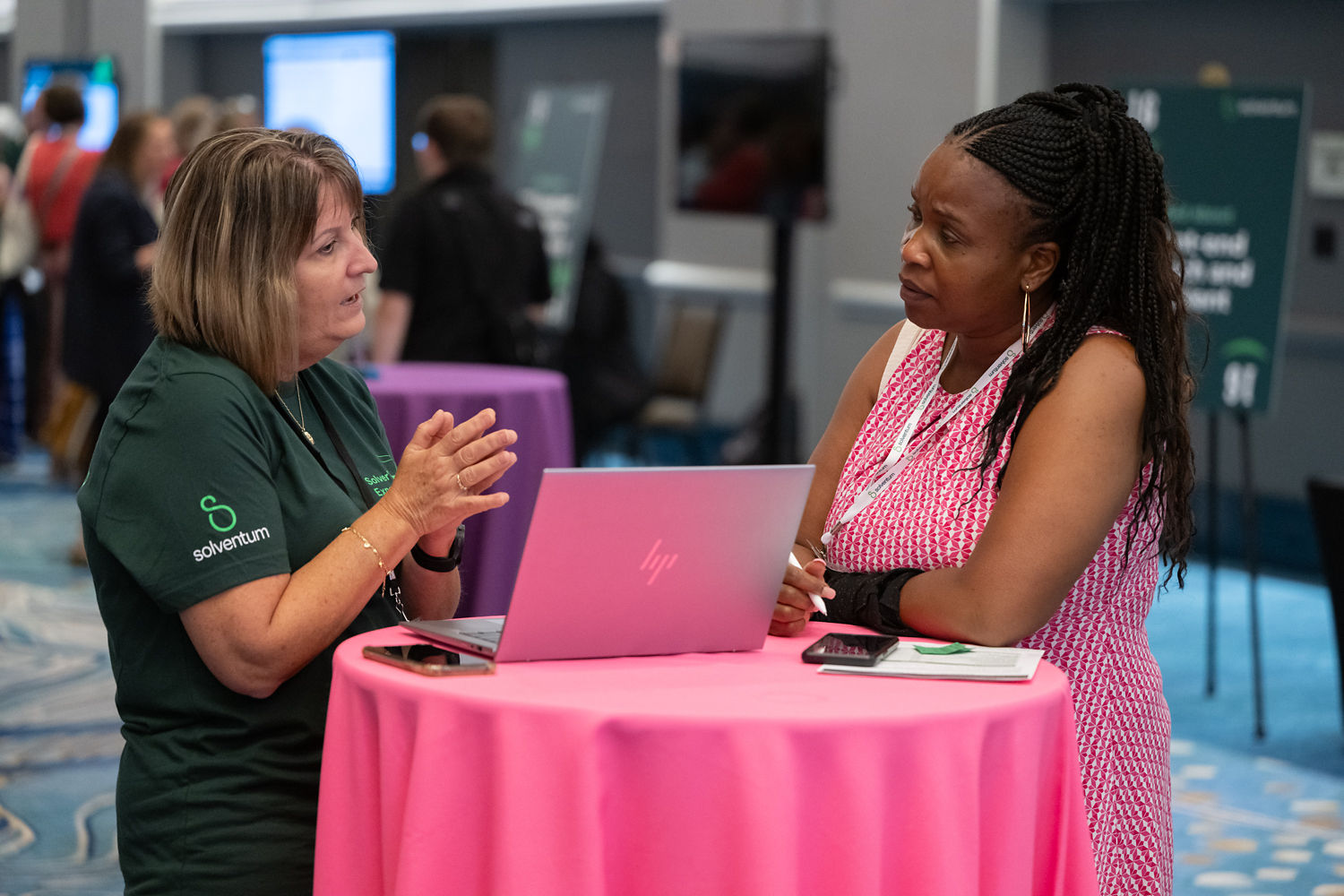 Two women standing at pink table with laptop, in conversation.