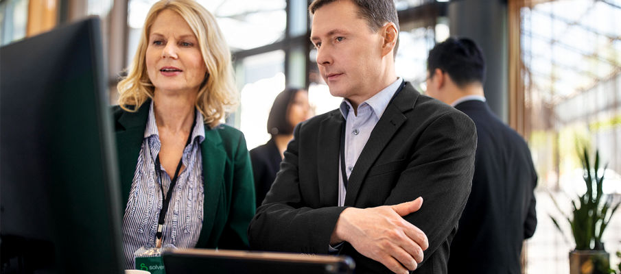 A female executive explaining her ideas to a businessman at a conference.  The female has blonde, shoulder length hair and is wearing a business professional green cardigan. The man has short brown hair and is wearing a business professional black suit coat. They are looking at a monitor.
