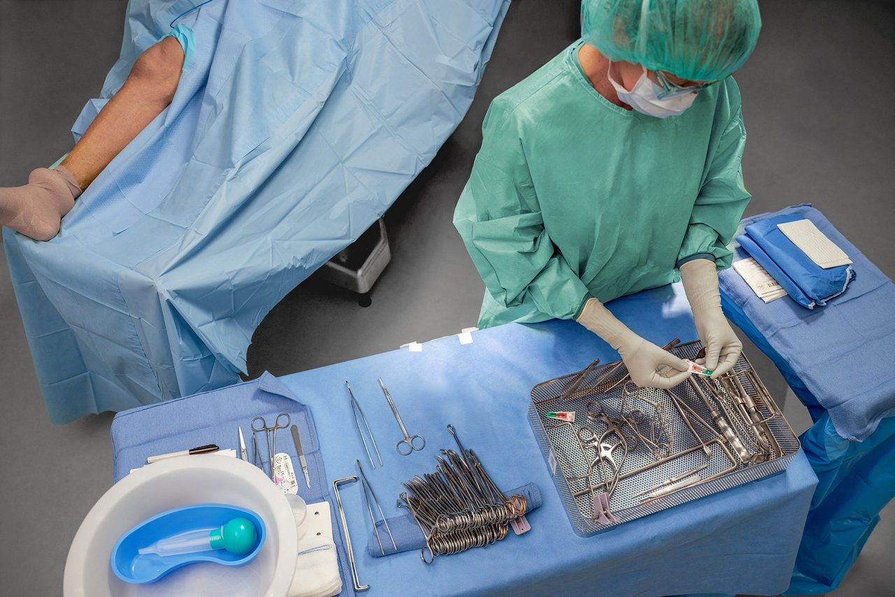 A clinician reviews a steam chemical integrator in a tray of instruments in the operating room. In the background, a patient's leg is visible and prepared for surgery.