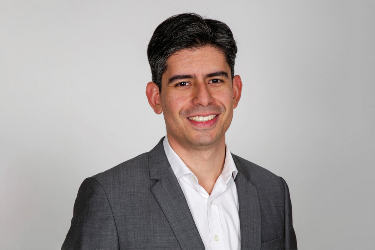 Un hombre sonriendo a la cámara, con una chaqueta de traje gris oscuro y camisa blanca, sobre un fondo gris claro.