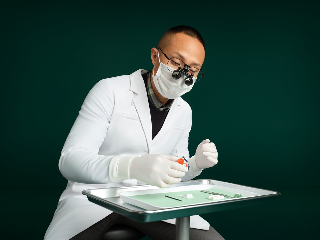 Dentista maschio seduto dietro un portafoglio dentale su un soporte che utilizza Scotchbond™ Universel Plus Adhesive, sfondo verde scuro.