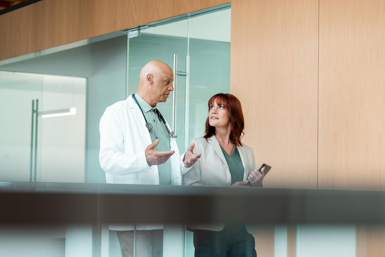 A male doctor, wearing a lab coat and Littman stethescope, walks alongside and talks to a female admin holding a tablet.