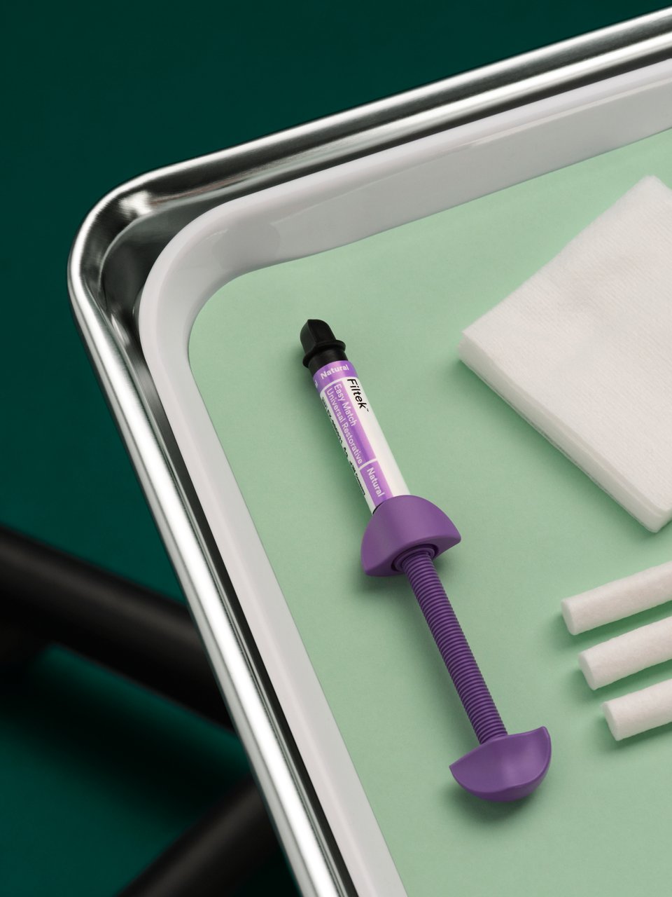 La jeringa restauradora universal 3M Filtek Easy Match se encuentra sobre una bandeja de metal para dentista con un inserto de plástico blanco y un revestimiento de color verde claro, el fondo del suelo es de color verde oscuro.