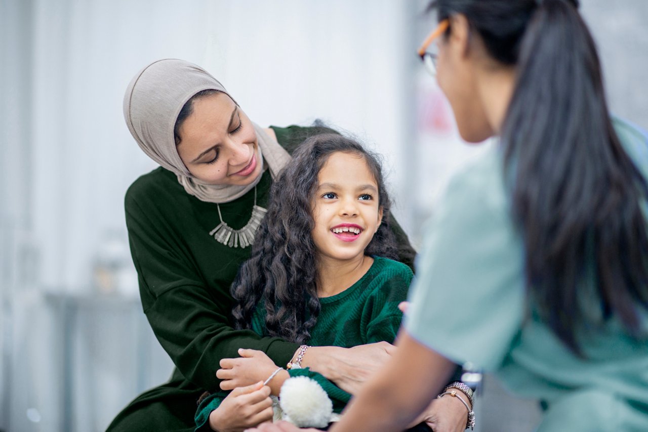 A smiling girl sits on a woman's lap while a female healthcare professional in scrubs interacts with them in a clinical setting.