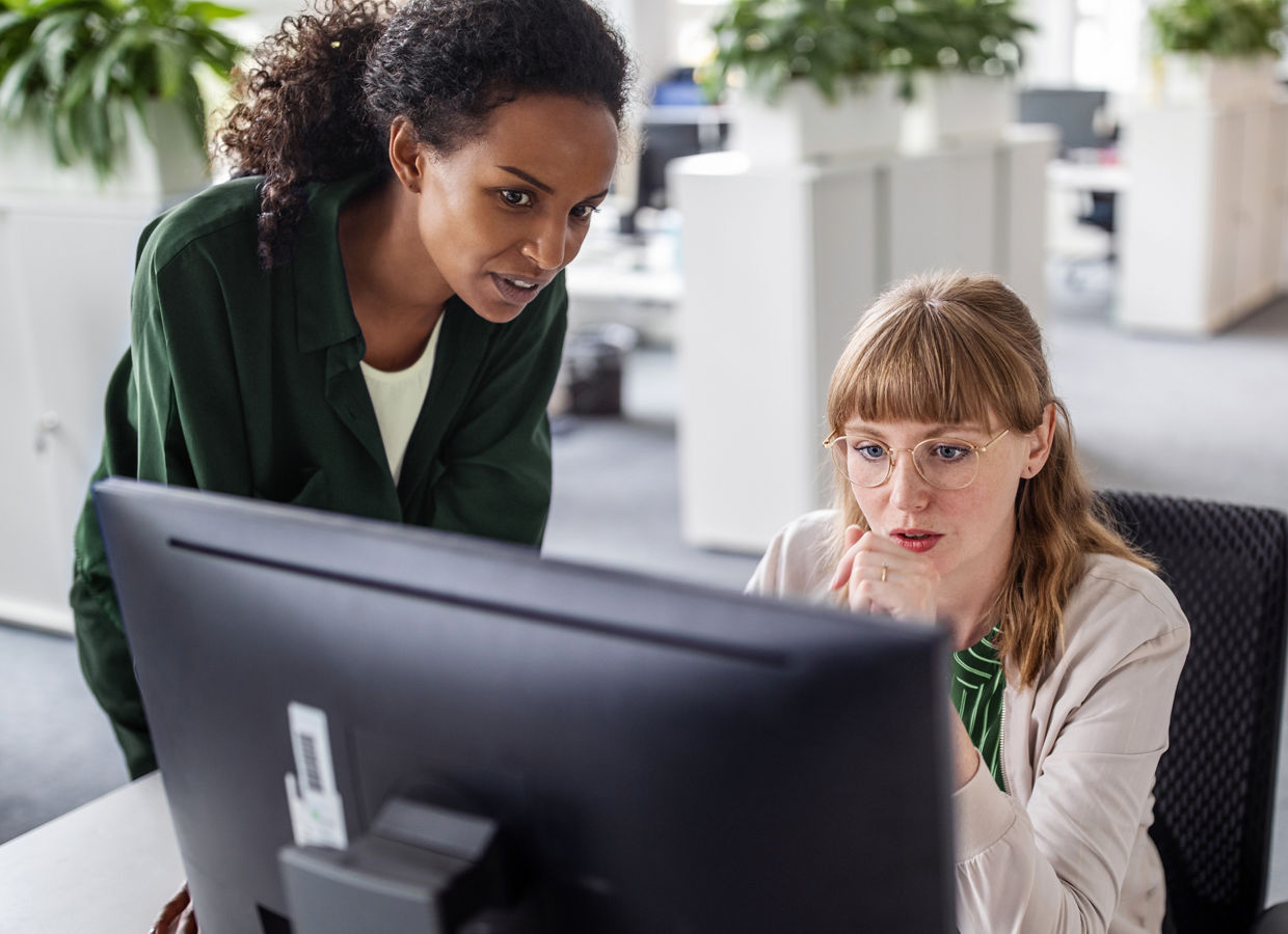 Two female professionals in an office collaborate while viewing a computer screen, with office furniture and plants in the background.
