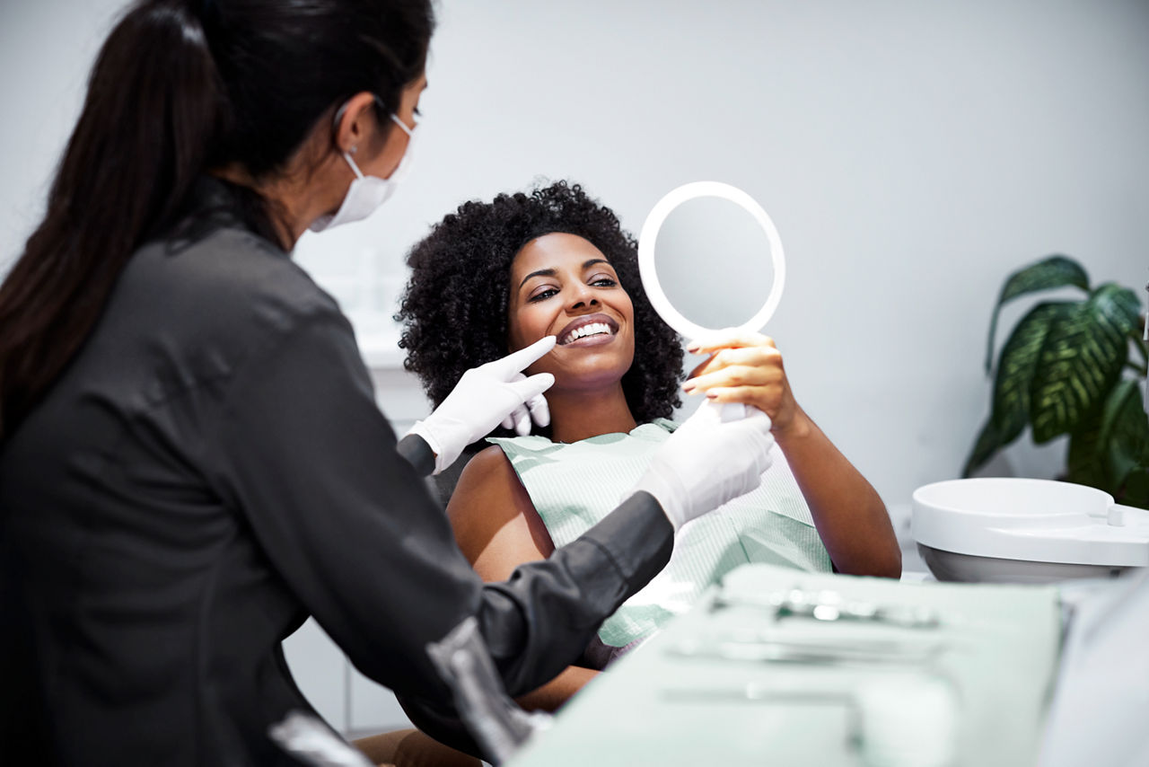 Smiling woman looking teeth in hand mirror. Female dentist is explaining dental health to patient. They are in medical examination room.