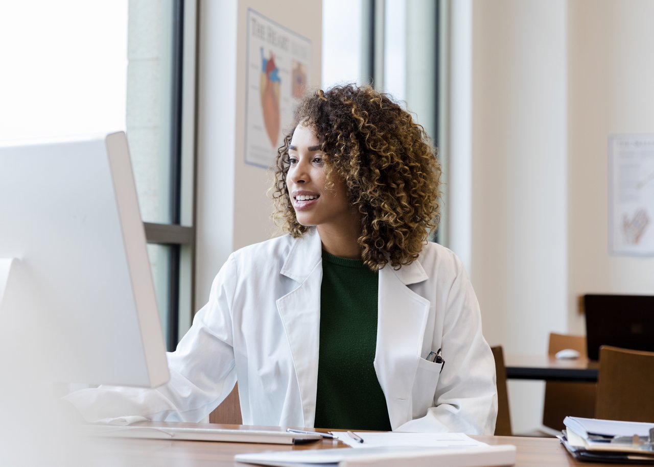 Mid adult female doctor in a white lab coat with a stethoscope, sits at a desk and reviews patient records on a computer.
