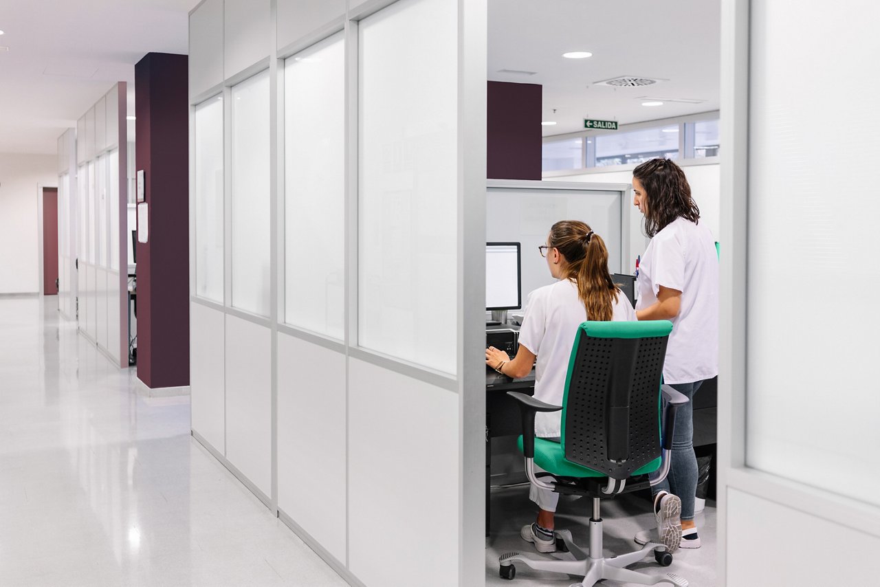 Two females in white uniforms are seen through a doorway working at a computer station in a modern office or medical setting with a white hallway.