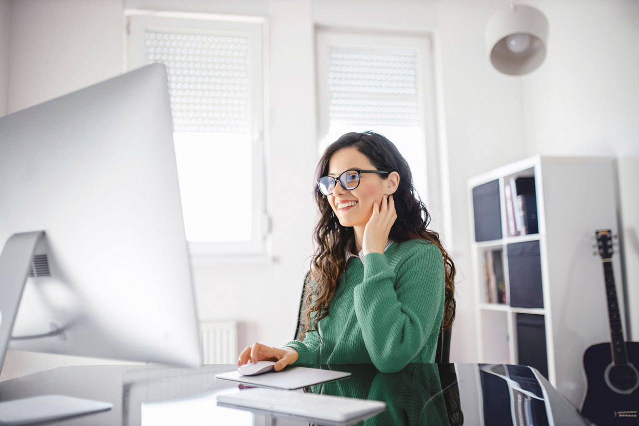 Smiling young woman in glasses and green sweater working at her home office desk with a large computer monitor.
