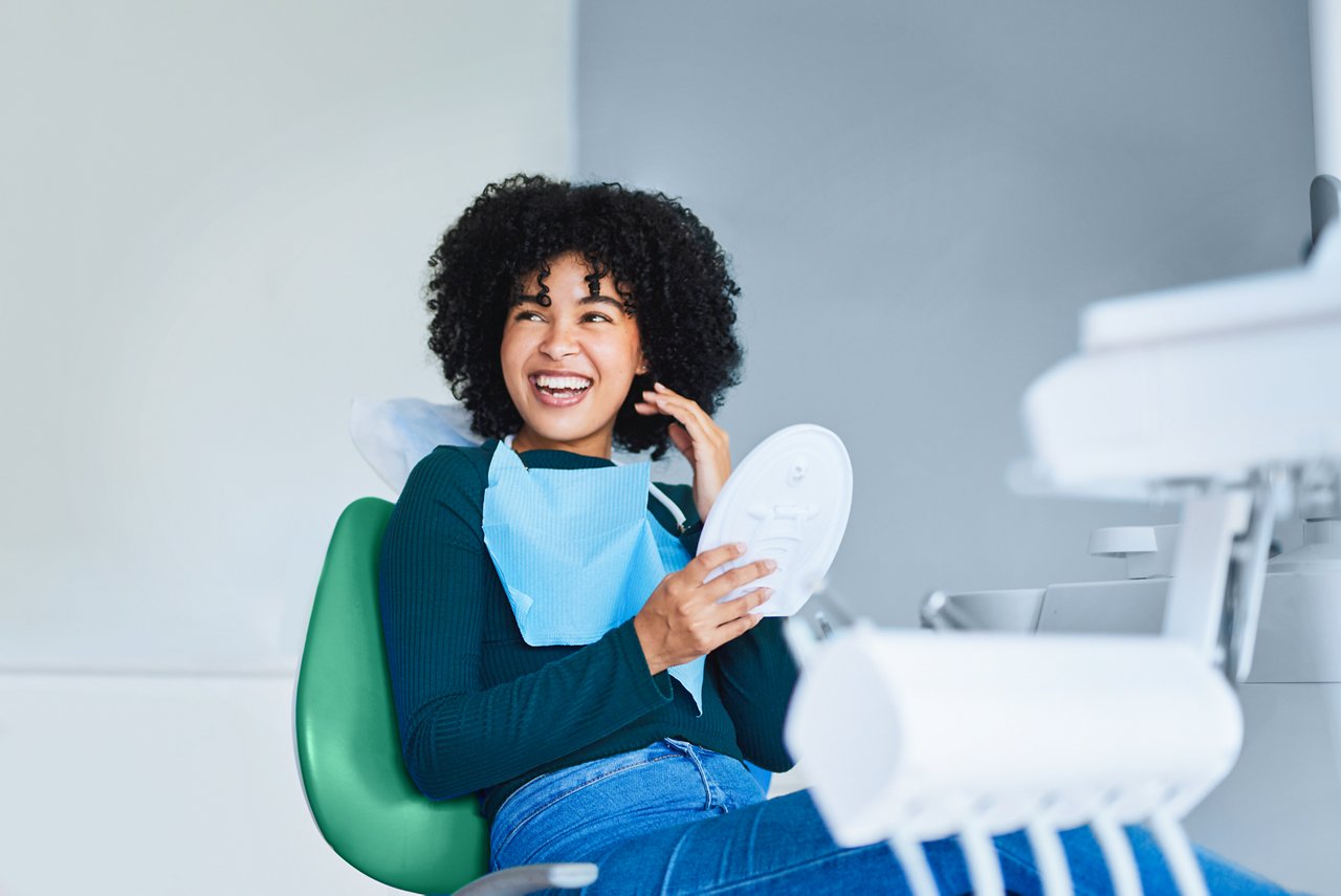 Un paciente adolescente en un sillón dental se ríe, mostrando unos dientes bonitos, mientras sostiene un espejo.