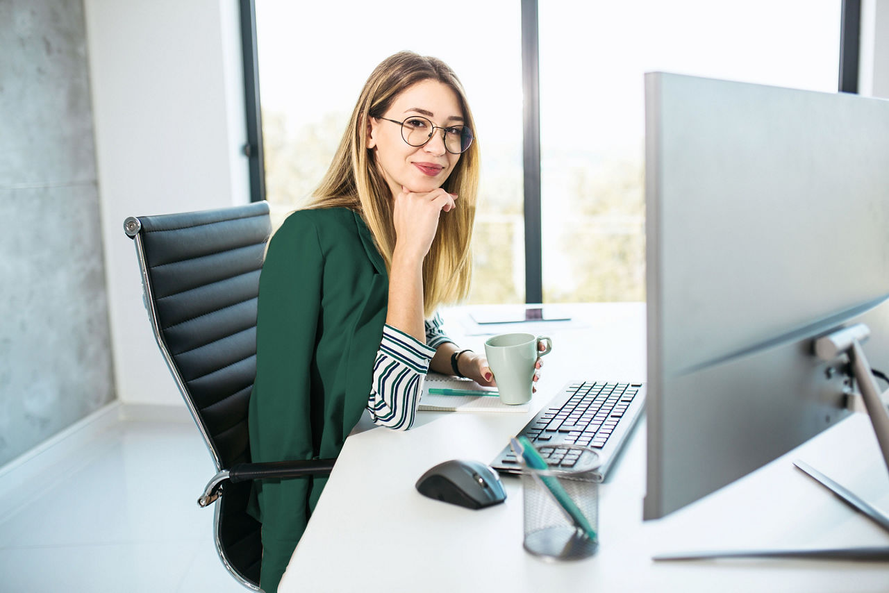 Smiling young female sitting at desk in a modern office, wearing a green blazer, holding a coffee mug, and looking at a computer monitor with a keyboard, mouse, and stationery on the desk.