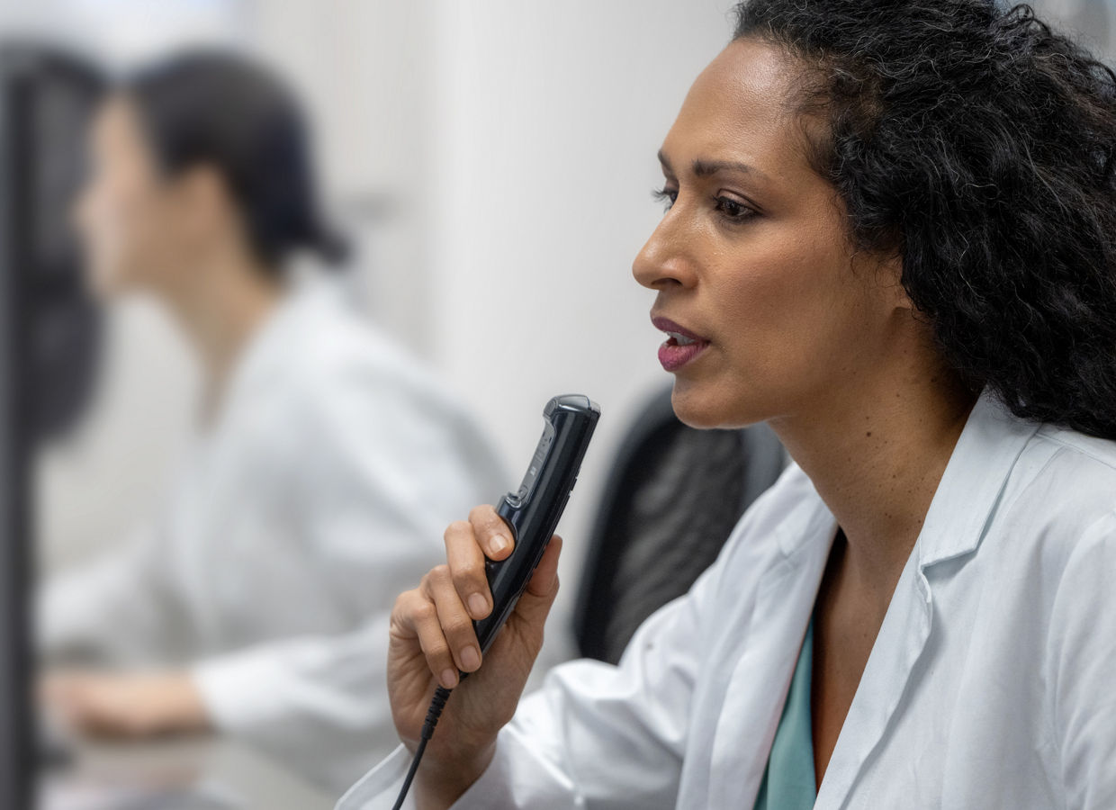 Female physician or doctor wearing a white lab coat, speaking into dictation recorder while looking at computer monitor, another person working at a computer in the background.