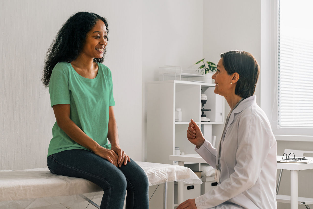 Cheerful and smiling African-American female patient talking to female doctor in exam room during appointment in medical clinic.
