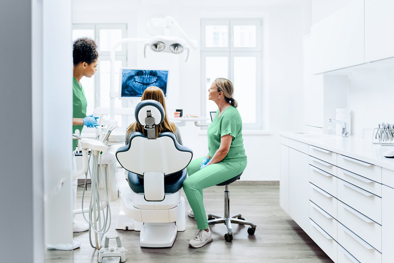 A female dentist seated on a stool consulting with a female patient in the dental chair. A female dentist assistant is standing next to the patient. All of them are viewing an x-ray on a monitor.