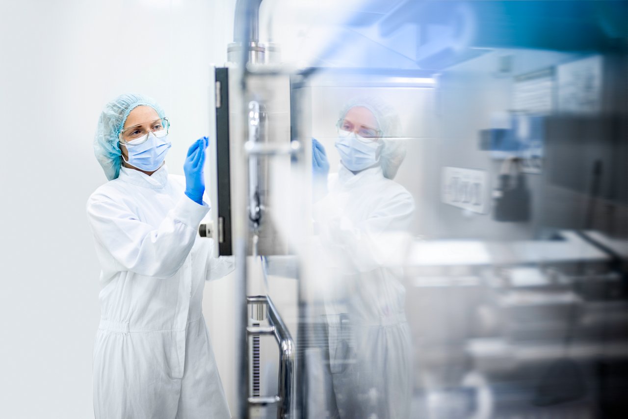 A woman wearing proper safety clothes seen taping on the screen of a big complex machine in pharmaceutical industry while her reflection is visible on the machine.