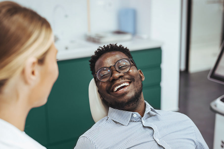 Smiling african guy in dentist chair looking with trust at his doctor, close up. Young African American Man Getting Teeth Treatment With Professional Stomatologist At Modern Clinic