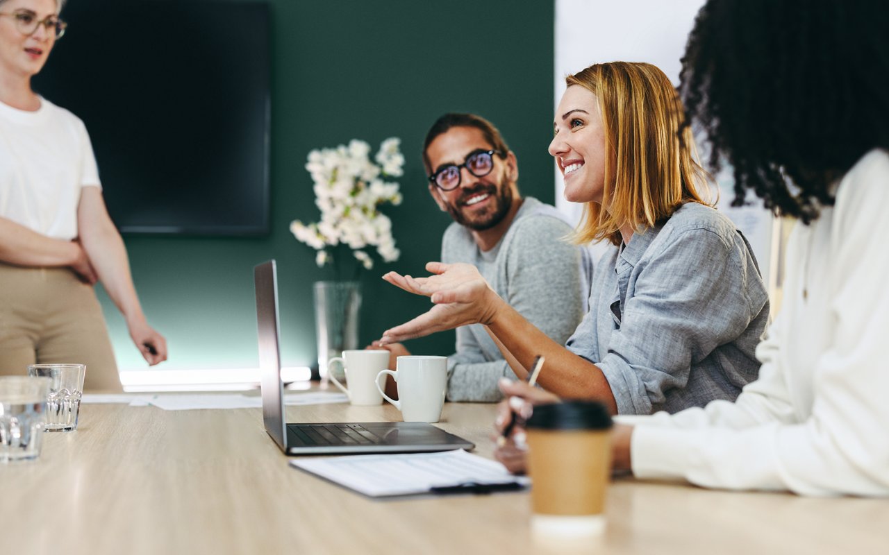 Varios colegas sonrientes conversan en una sala de conferencias. Uno de ellos está de pie y los demás están sentados en la mesa de conferencias.