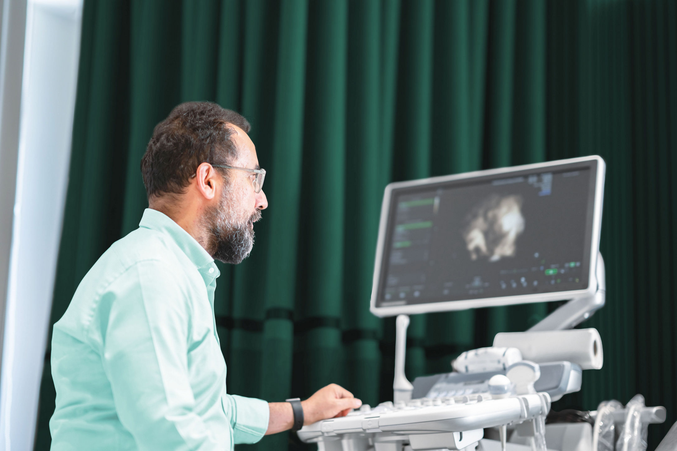 Male obstetrician-gynecologist doctor in medical examination room reviewing an ultrasound on equipment.