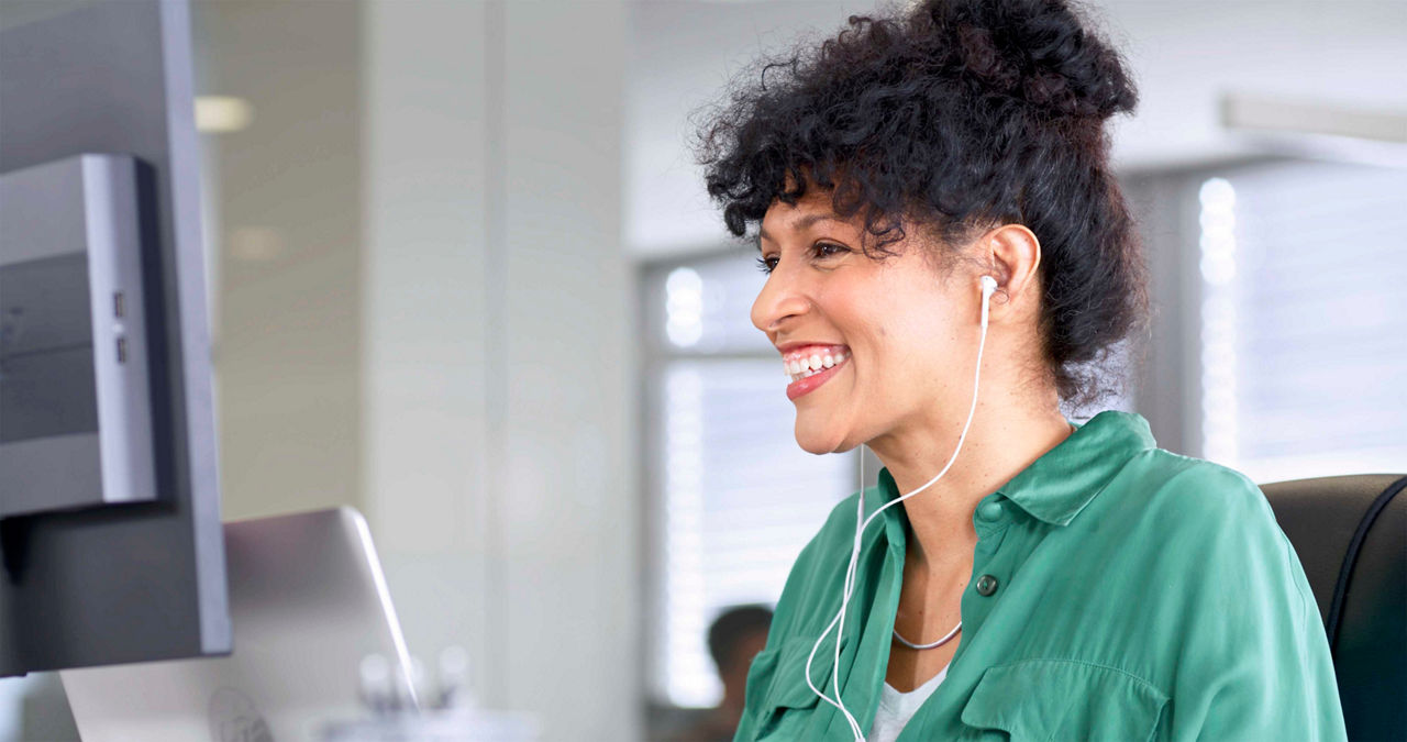 A smiling businesswoman wearing earphones participates in a computer conference call while sitting in a modern office.