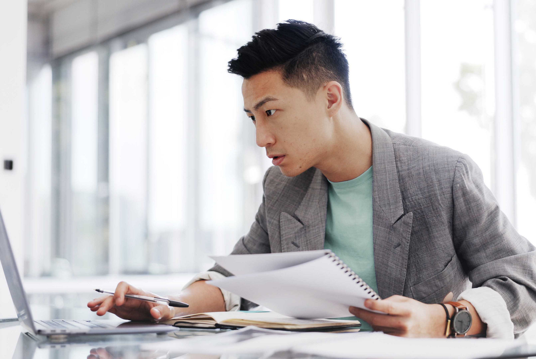 An Asian businessman works in his office, writing reports and business plan documents on his laptop while taking notes with a pen for development, audit, and strategy.