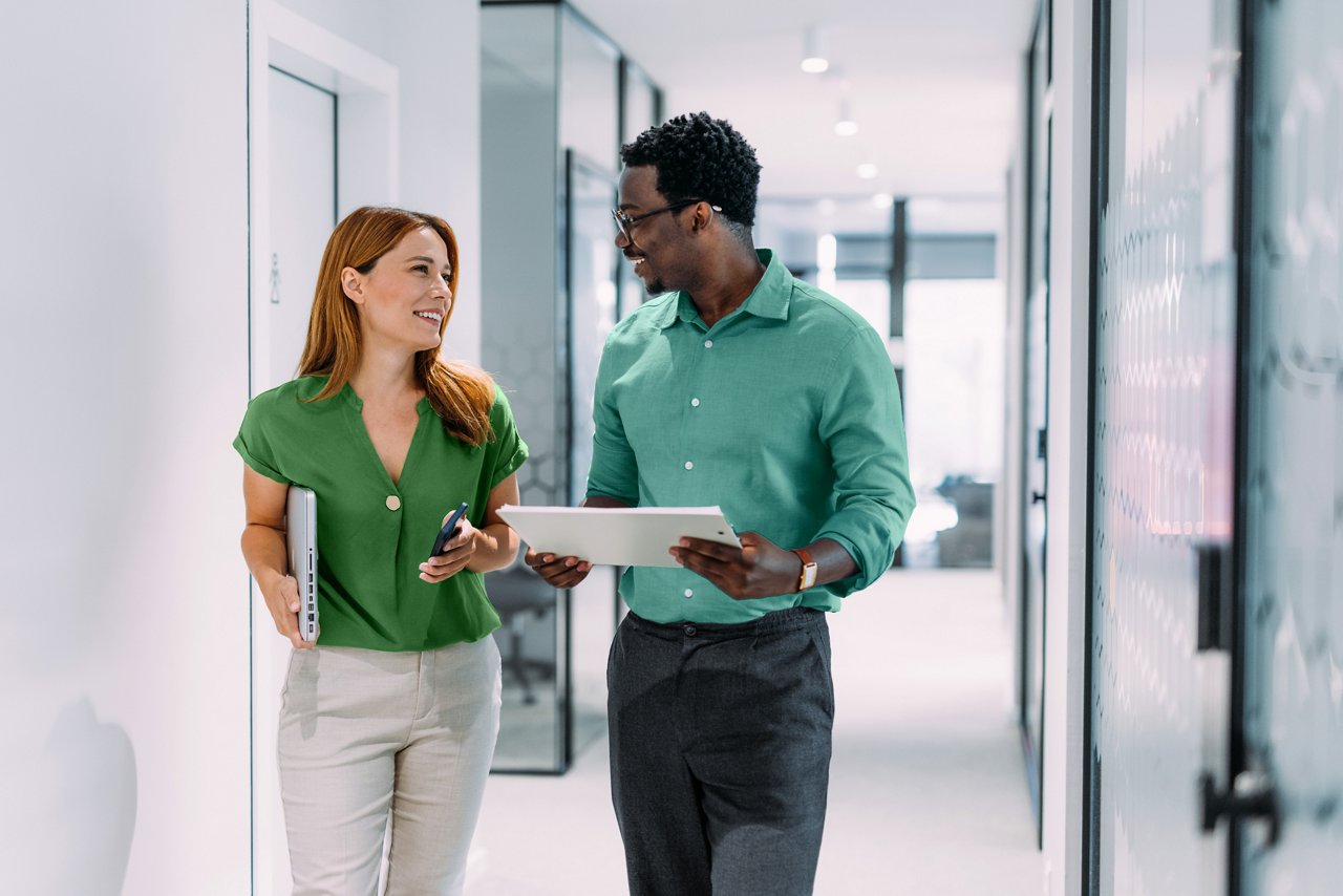 Un uomo e una donna d'affari sicuri di sé camminano attraverso il corridoio di un moderno ufficio, tenendo un laptop, uno smartphone e un tablet, impegnati in conversazione.