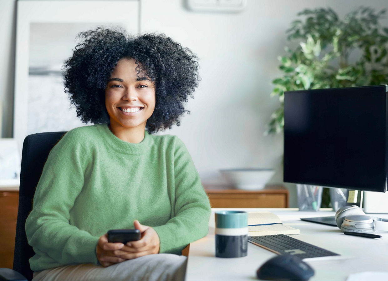 A smiling young woman sits at her desk holding a phone in her home office.
