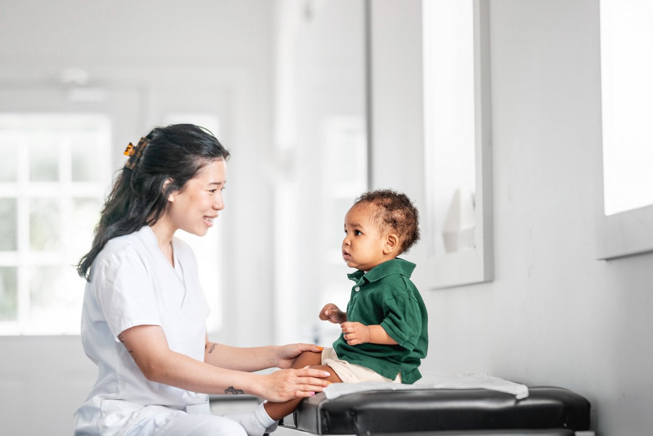 Asian female pediatrician interacting with curious toddler during check-up in bright clinic room.