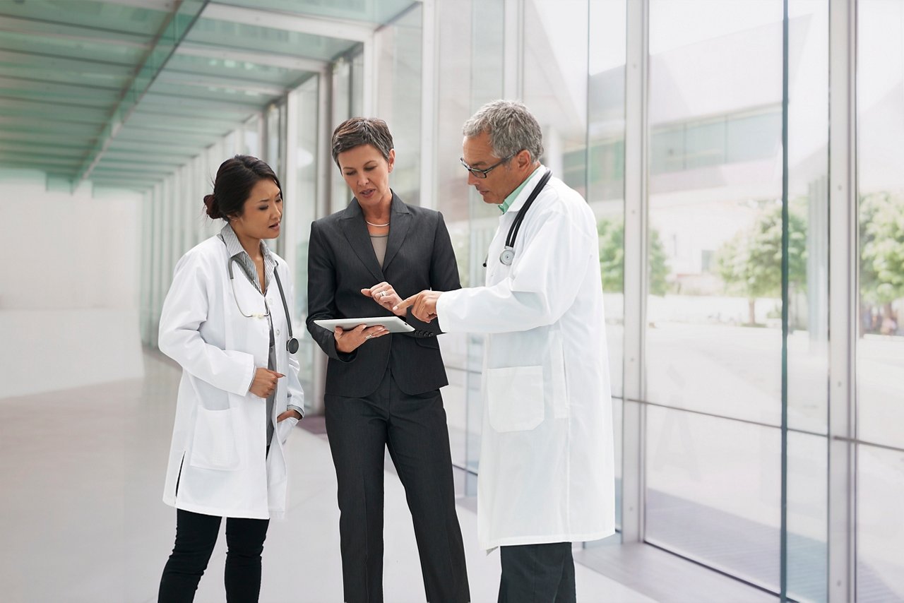 Three medical professionals meet in a bright, glass-walled corridor, discussing something on a tablet.