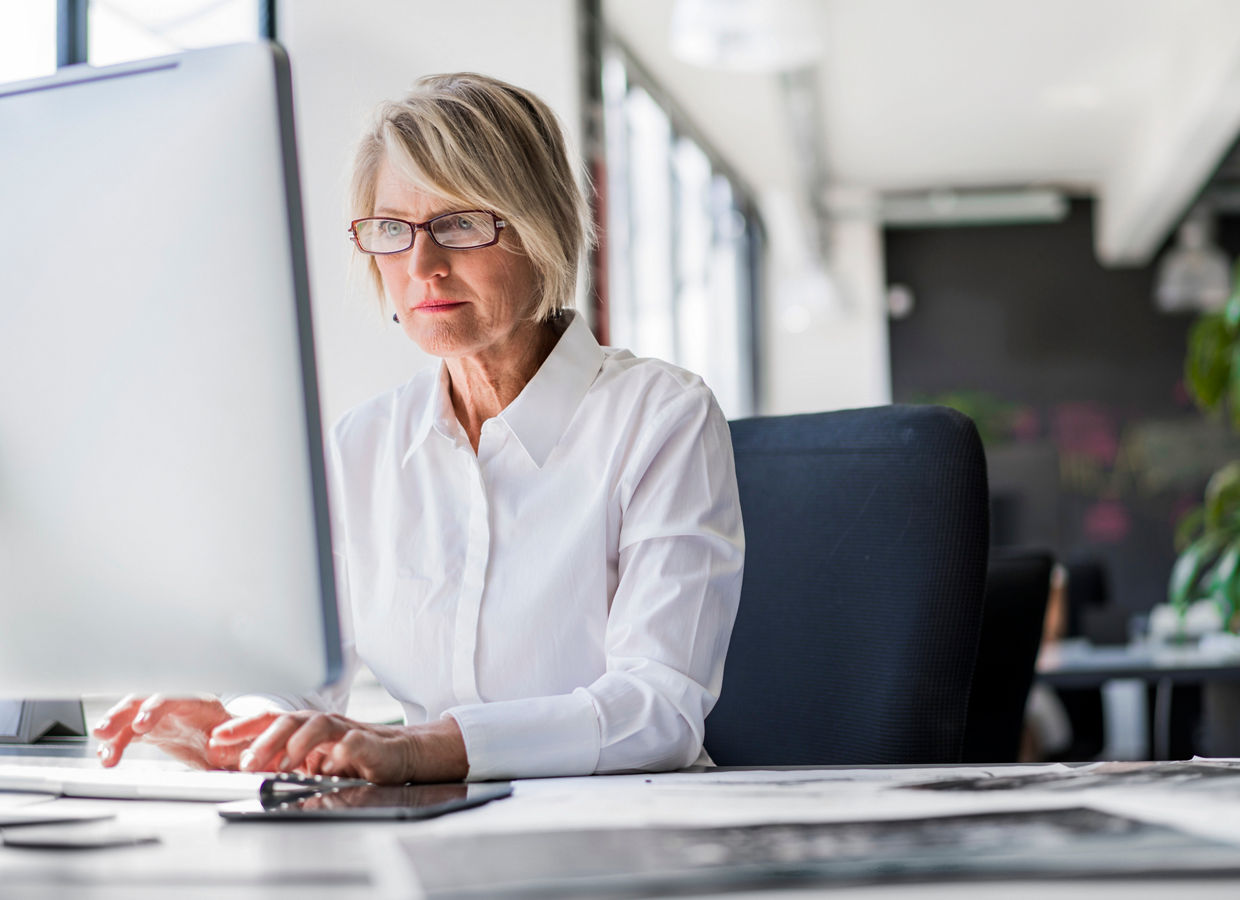 Mature businesswoman in glasses and white shirt working at a desk computer in a bright office with large windows and plants.