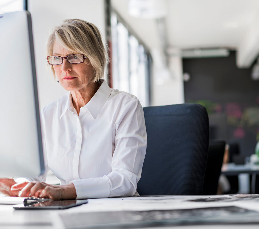 Mature businesswoman in glasses and white shirt working at a desk computer in a bright office with large windows and plants.