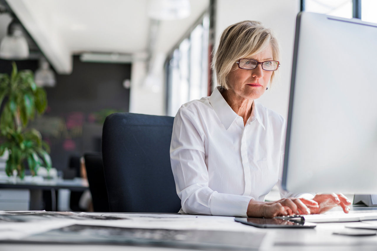 Mature businesswoman in glasses and white shirt working at a desk computer in a bright office with large windows and plants.