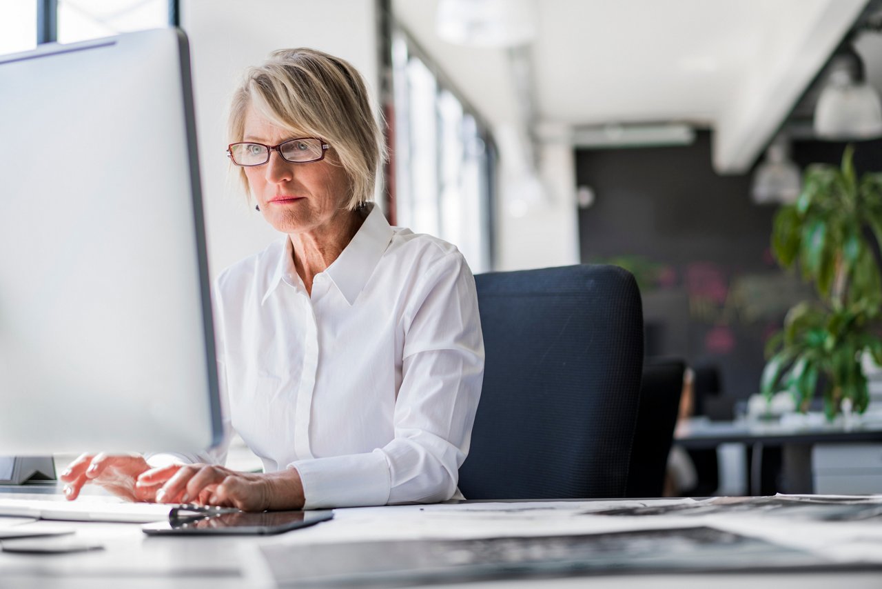 Mature businesswoman in glasses and white shirt working at a desk computer in a bright office with large windows and plants.
