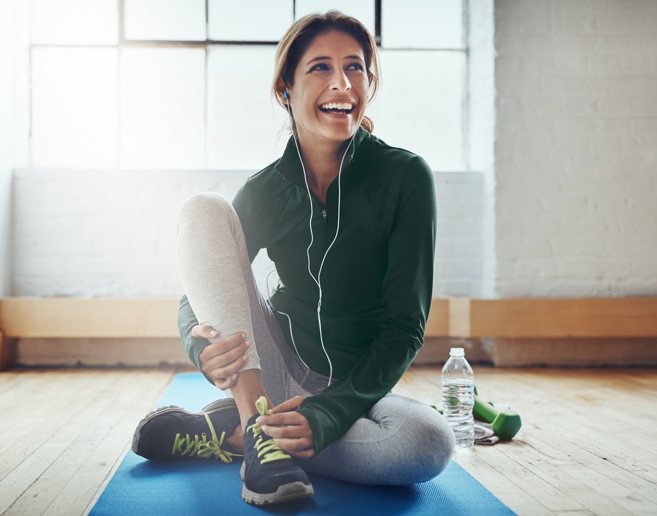 Smiling woman sitting on a yoga mat indoors, wearing workout clothes and earphones, with a water bottle and dumbbell nearby.