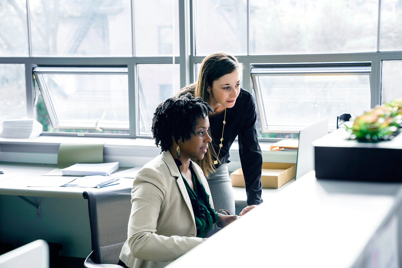 Two businesswomen collaborate at a desk in a sunlit office, focused on a computer screen with papers and supplies nearby.