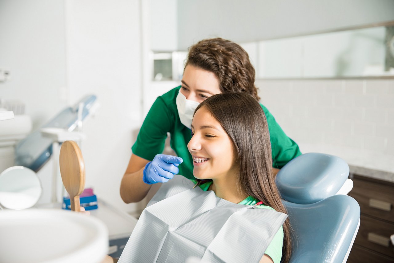 In scrubs by a tray table, a seated orthodontist smiles at the camera.