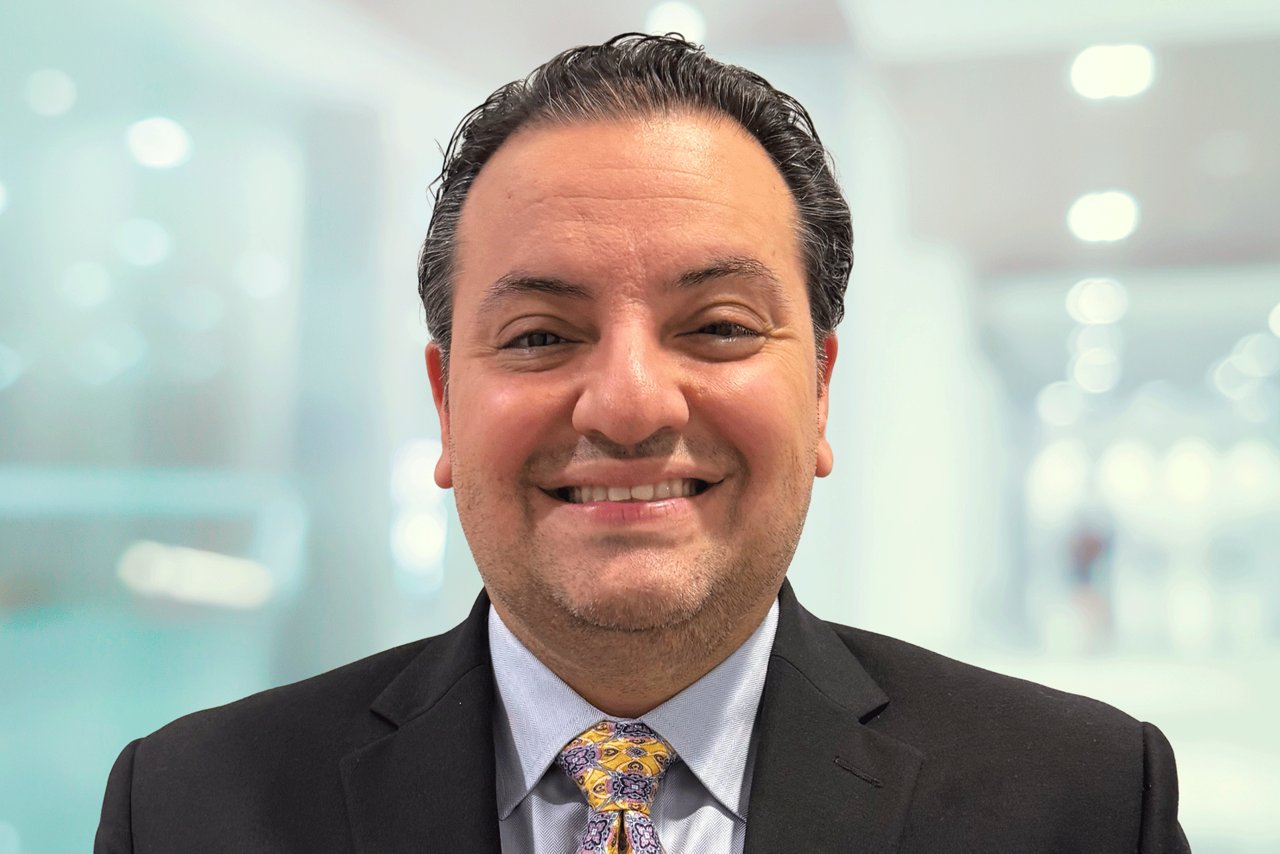 Headshot of a smiling man in a black blazer, light colored shirt and yellow tie in an office setting.