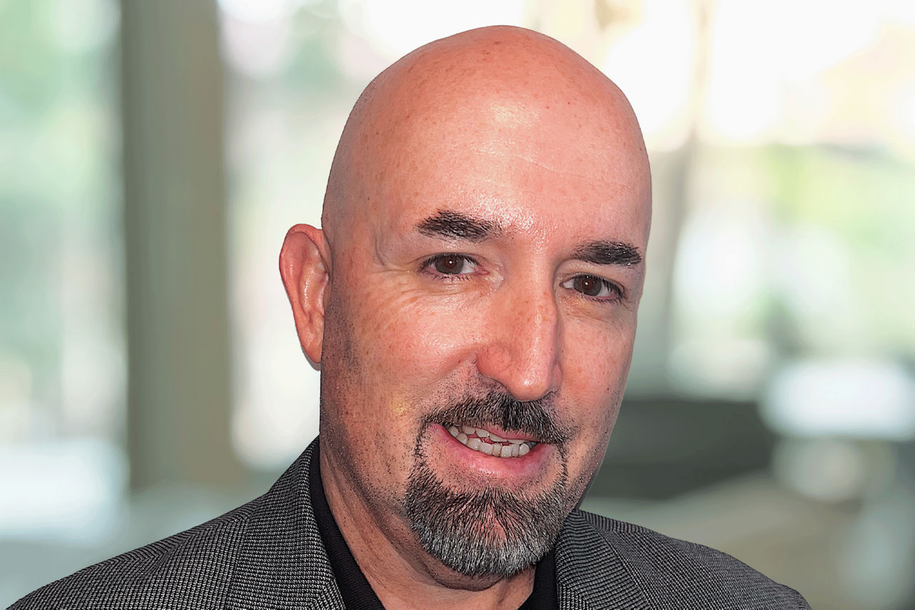 Headshot of Ed Lowry smiling and wearing a professional shirt and black jacket against and office background.