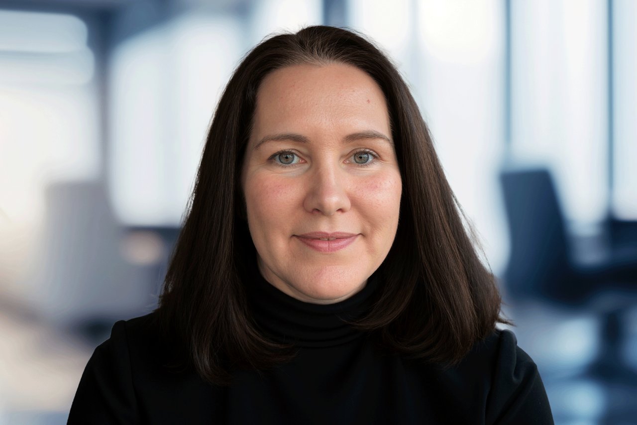 A woman with dark brown hair and black shirt, smiling at camera with an office setting background.