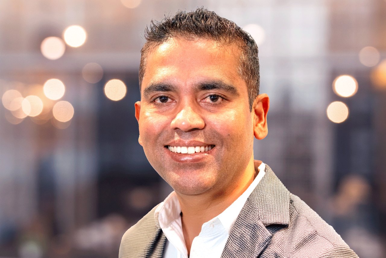Headshot of Hari Bala smiling and wearing a professional shirt and black jacket against and office background.