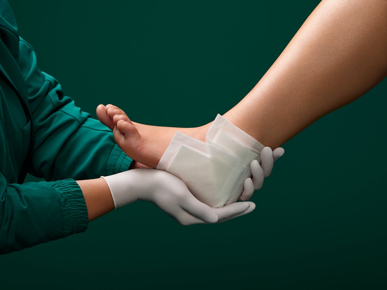 Portrait of a female nurse holding a Kerramax Care Super-Absorbent Dressing onto the heal of a wound care patient on a dark green background.