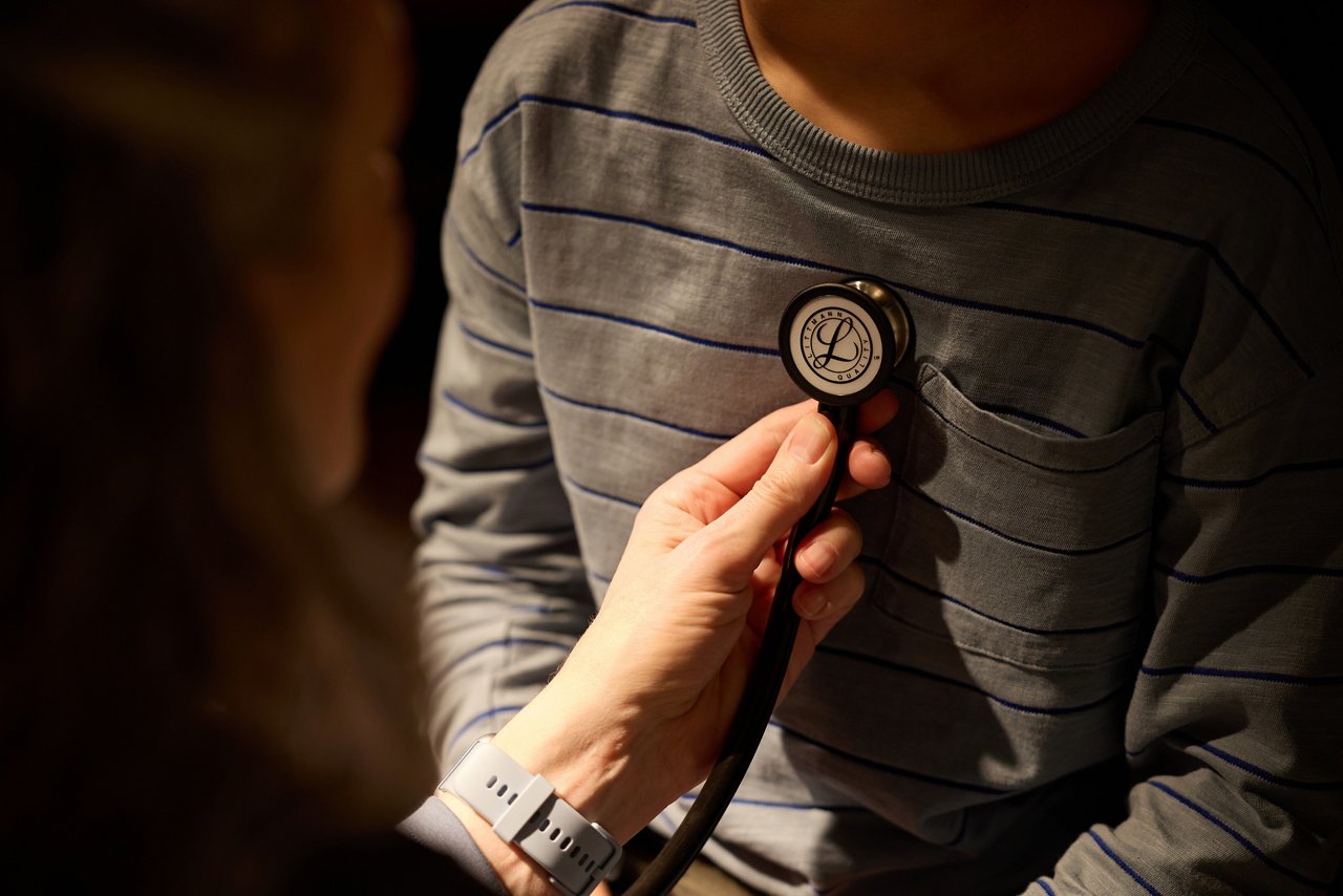 A close-up view shows a healthcare professional placing a Littmann stethoscope chestpiece against a child patient’s shirt during a medical examination.