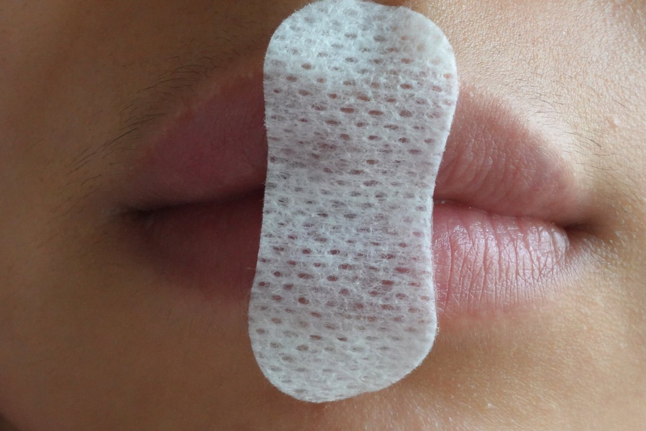 A white textured adhesive mouth tape strip is placed vertically over closed lips for application.