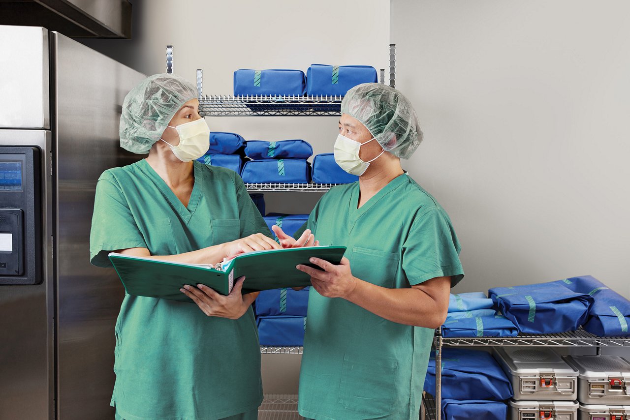 One female and one male technician in central sterile service  department holding  an open binder in front of shelves next to sterilizer